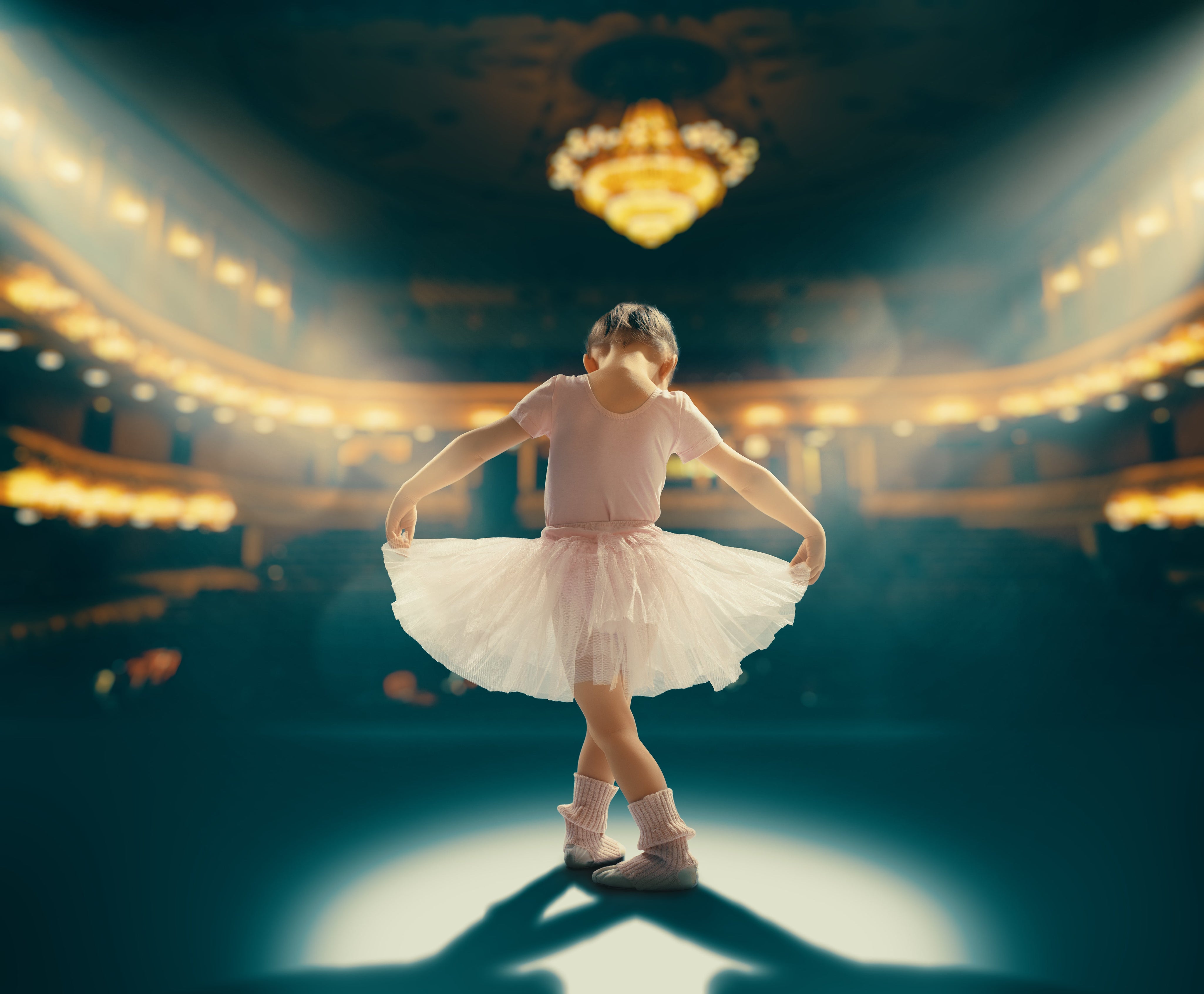Dance Recitals: Young ballerina in pink leotard and white tutu standing in spotlight on stage, performing with elegant theater backdrop and chandelier